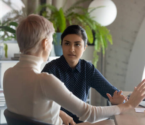 Indian and caucasian businesswomen negotiating sit at desk in office. Lawyer consulting client during formal meeting. Job interview and hiring process. Diverse colleagues discussing project concept