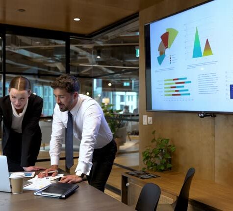 Business man and woman meeting and talking. There is paperwork and technology on the table and a TV in the background with financial graphs charts and figures.